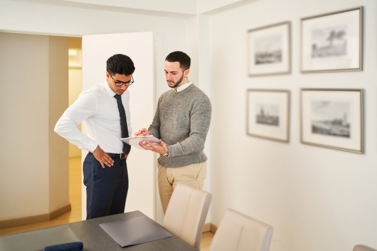 Two men are standing in front of a table with papers on it. One of them is wearing a tie